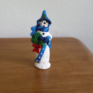 May include: A white snowman figurine wearing a blue hat and scarf, holding a green wreath with a red bow. The snowman has black eyes and a black mouth.