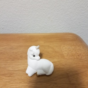 May include: A white ceramic unicorn figurine with a gold horn and hooves. The unicorn is sitting on its hind legs with its head turned to the side.