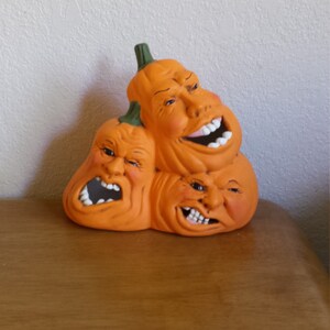 Könnte beinhalten: Drei Keramik-Kürbisse mit verschiedenen Gesichtsausdrücken. Die Kürbisse sind übereinander gestapelt und haben eine leuchtend orange Farbe.