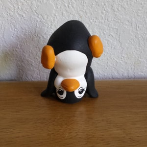 May include: A ceramic penguin ornament in a playful pose. The penguin is black with a white belly and face, and orange accents on its ears and beak. The eyes have delicate eyelashes. The ornament is on a wooden surface.