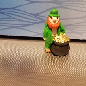 May include: A small, hand-painted figurine of a leprechaun in a green outfit and hat, with an orange beard, smiling and standing next to a black pot overflowing with gold coins. The figurine is on a textured surface.