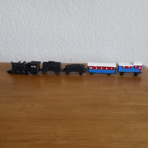 May include: A miniature toy train set featuring a black locomotive, two black cargo cars, and two passenger cars with red, white, and blue accents. The train is displayed on a wooden surface.