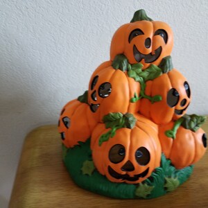 Könnte beinhalten: Ein Stapel Keramik-Kürbisse mit geschnitzten Gesichtern, angeordnet in Pyramidenform. Die Kürbisse sind orange mit schwarzen Augen, Nasen und Mündern. Die Kürbisse sind von grünen Blättern umgeben.