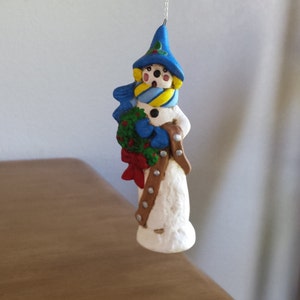 May include: A snowman ornament with a blue hat, yellow scarf, and a red bow holding a green wreath.