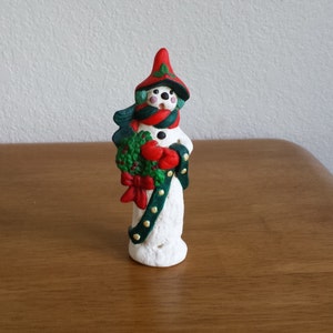Könnte beinhalten: Eine weiße Schneemann-Figur mit rotem Hut, grünem Schal und einem grünen und roten Kranz in den Armen. Der Schneemann ist mit goldenen Punkten verziert.