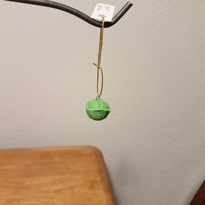 May include: A green, textured, round ornament hangs from a gold string attached to a metal hook. The ornament is labeled "$2.50".