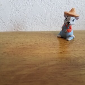 May include: A small ceramic figurine of a mouse wearing overalls and a straw hat. The mouse is sitting on a wooden surface.