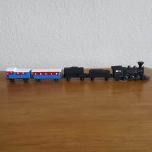 May include: A toy train set with two blue passenger cars, a black coal car, a black flat car, and a black steam engine. The train is on a wooden surface.
