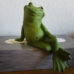 May include: A green frog figurine sitting on a gray tissue box with a white tissue insert. The frog is sitting with its legs crossed and its arms resting on its legs. The frog is looking up.