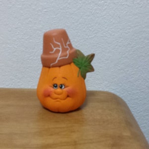May include: A ceramic pumpkin figurine with a brown pot on its head and a green leaf. The pumpkin has a painted face with a smile and closed eyes.