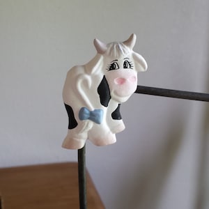 May include: A white and black ceramic cow figurine with a blue bow, mounted on a black metal stand. The cow has a pink nose and black eyes.
