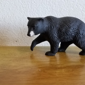 Pode incluir: Uma figura de urso preto com marcas brancas ao redor dos olhos. O urso está andando com a cabeça baixa e suas patas são visíveis. A figura é feita de um material escuro e texturizado e está em pé sobre uma superfície de madeira.