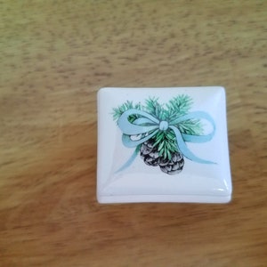 May include: A white square trinket box with a blue ribbon and pine cone design.