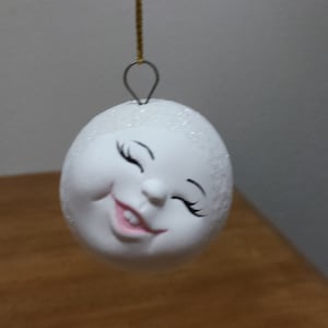May include: White ceramic ornament shaped like a face with a smiling expression and closed eyes. The ornament is hanging from a gold string.