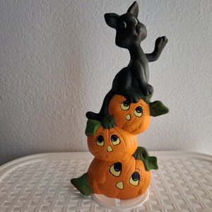 Könnte beinhalten: Eine schwarze Keramik-Katzenfigur, die auf einem Stapel von drei orangefarbenen Keramik-Kürbissen mit bemalten Gesichtern sitzt. Die Kürbisse sind mit grünen Blättern verziert.