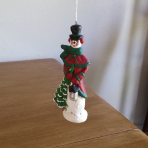 Op de afbeelding: Een sneeuwman ornament met een rode en groene hoed, sjaal en jas, die een lantaarn vasthoudt. De sneeuwman staat op een witte basis.