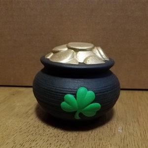 May include: A black ceramic pot with a gold-colored lid and gold coins. A green shamrock is on the front of the pot. The pot is a St. Patrick's Day decoration.