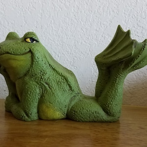 Könnte beinhalten: Grüne Keramik-Froschfigur mit einem skurrilen Ausdruck, sitzend mit ausgestreckten Beinen und einem eingerollten Schwanz.