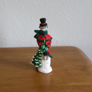 Op de afbeelding: Een wit sneeuwpopfiguur met een zwarte hoge hoed, een groene sjaal en een rode jas. De sneeuwpop houdt een kleine groene kerstboom vast.