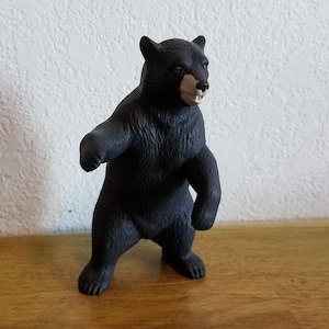 May include: A black bear figurine in a standing pose. The bear has a detailed fur texture and a light brown snout. The figurine is on a wooden surface with a white textured wall in the background.