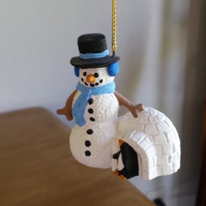 May include: A snowman Christmas ornament with a black top hat, blue earmuffs, and a blue scarf. The snowman is holding a white igloo with a penguin inside. The ornament hangs from a gold string.