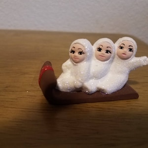 May include: Three white snowmen figurines with black eyes and eyelashes riding in a brown sled with red accents.