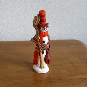May include: A whimsical snowman figurine with a red and brown hat, a red scarf, and a brown bag. The snowman is holding a broom in one hand and a brown bag in the other.