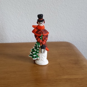 May include: A small snowman figurine with a black top hat, red scarf, and green tree in hand. The snowman is made of white ceramic and has a brown coat.