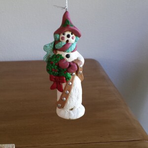 Puede incluir: Un adorno de muñeco de nieve blanco con un sombrero rojo, acebo verde y un cinturón marrón. El muñeco de nieve sostiene una manopla roja y lleva una bufanda azul.
