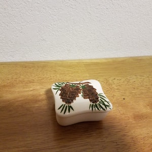 May include: A white ceramic trinket box with a square shape and a slightly curved lid. The lid features a painted design of two pine cones with green needles on a branch.