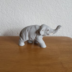 May include: A gray ceramic elephant figurine with its trunk raised. The elephant is standing on a wooden surface.
