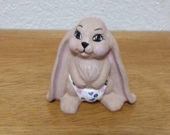 Könnte beinhalten: Keramik-Hasenfigur mit langen, schlaffen Ohren und einem süßen Ausdruck. Der Hase ist hellbraun mit rosa Akzenten an den Ohren und einem Höschen mit Herzmuster. Die Figur steht auf einer Holzoberfläche.
