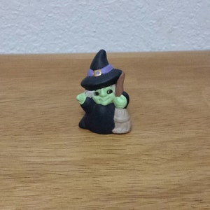 Puede incluir: Una pequeña figura de bruja verde con un sombrero negro y ribete morado, sosteniendo una escoba marrón.