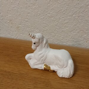 May include: A white ceramic unicorn figurine with a gold horn and hooves. The unicorn is lying down with its head turned to the side.