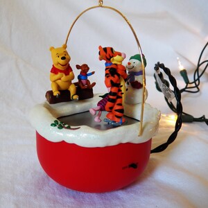 Peut inclure: Une décoration de Noël musicale rouge et blanche représentant Winnie l'ourson, Tigrou, Porcinet et un bonhomme de neige faisant du patin à glace. La décoration est dotée d'un cadre en fil de fer doré et d'une guirlande lumineuse à piles.