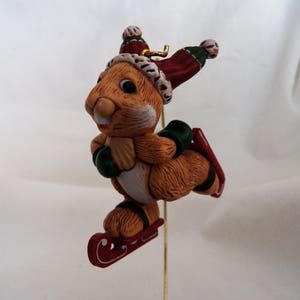 Peut inclure: Une décoration de Noël en céramique marron et blanche représentant un écureuil portant un bonnet de Père Noël rouge et blanc et des mitaines vertes, faisant du patin à glace sur un traîneau rouge.