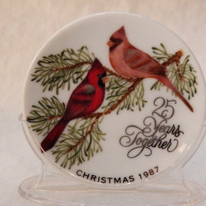 Puede incluir: Un adorno navideño de cerámica blanca con dos cardenales rojos posados en una rama de pino. El adorno dice "25 Years Together" y "Christmas 1987".