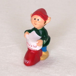 May include: A small figurine of an elf wearing a red hat, green shirt, and blue pants, holding a white stocking with the red text Baby's 1st Christmas. The red stocking features the white text 1989 on the toe, and the elf wears yellow shoes.