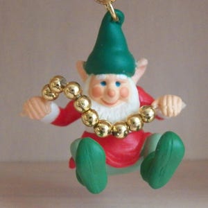 May include: A Christmas ornament featuring a seated elf with a red coat, green hat and shoes, and a white beard. The elf is wearing a gold beaded necklace and has rosy cheeks. A gold hook is at the top for hanging.