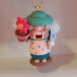 May include: A wooden nutcracker ornament featuring a figure in a green hat and pink polka-dot outfit. The figure holds a small terracotta-colored flower pot with a red flower and the year 1997 printed on it, along with a silver-colored garden trowel.