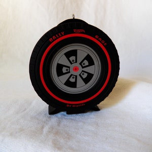 May include: Black and red Hot Wheels tire-shaped ornament. The tire has a red stripe and the words "RALLY" and "GASE" in red. The center of the wheel is gray with black accents. The ornament is made by Mattel.