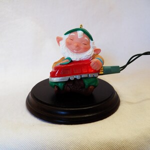 Könnte beinhalten: Ein grüner und roter Weihnachtsbaumschmuck, der einen Gnom zeigt, der ein rotes Spielzeug-Zugs spielt. Der Gnom trägt einen grünen Hut und hat einen weißen Bart. Der Schmuck befindet sich auf einem Holzsockel und hat einen Draht daran.