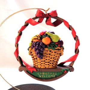 May include: A decorative hanging ornament featuring a woven basket filled with assorted fruits including purple grapes, a green pear, and an orange. A red ribbon is tied at the top, and a green plaque at the base displays the text CHRISTMAS WELCOME.
