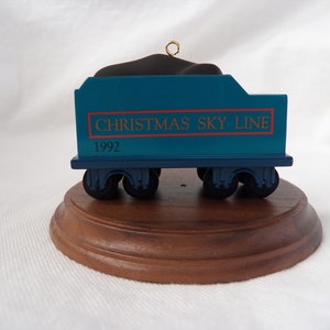 以下が含まれることがあります： 「CHRISTMAS SKY LINE」と「1992」の赤い文字が入った、水色のクリスマスの飾り。列車の車両の形をしており、木製の台座の上に置かれています。黒い車輪と黒い上部があります。