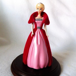 Peut inclure: Une figurine de poupée en porcelaine représentant une femme vêtue d'une cape en velours rouge et d'une robe en satin rose. Elle a les cheveux blonds et porte une tiare. La poupée est posée sur un socle en bois brun foncé.