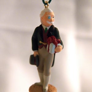 May include: A hanging ornament depicting a figure with white hair and a mustache, wearing a dark green coat, a maroon vest, and light tan trousers. The figure holds a white gift box with a red ribbon and a black top hat in the other hand.