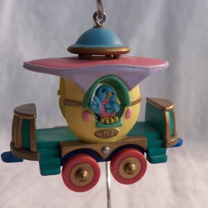 May include: A whimsical, colorful train ornament with a yellow egg-shaped car, a blue bird passenger, and a pink and purple roof. The train has gold accents and green wheels.