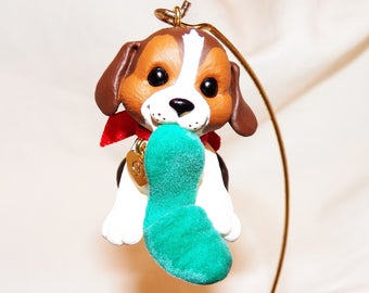 Könnte beinhalten: Ein brauner und weißer Beagle-Hunde-Anhänger mit einer roten Schleife und einem grünen Strumpf im Maul. Der Anhänger hängt an einem goldenen Haken.
