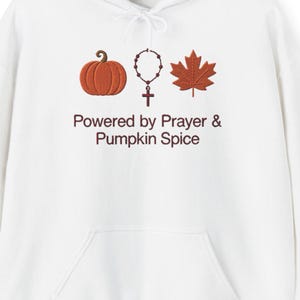 May include: A white hooded sweatshirt featuring a graphic print of an orange pumpkin, a brown rosary, and a brown maple leaf. Below the icons, the text reads "Powered by Prayer & Pumpkin Spice" in a dark brown, sans-serif font.