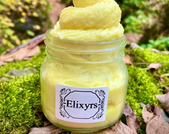 May include: A glass jar filled with a yellow, whipped body butter. The jar has a white label with the word "Elixyrs" in black lettering.